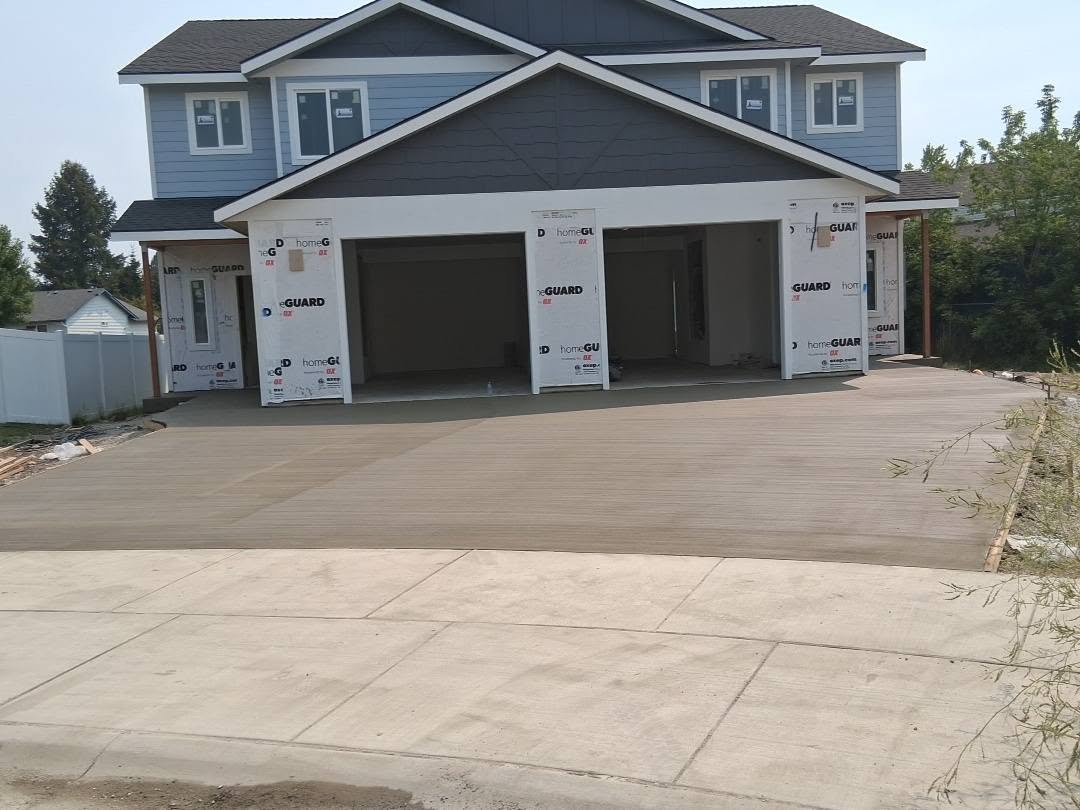 A blue-gray two-story house under construction, with open double garage bays and a broad concrete driveway in bright, quiet daylight.