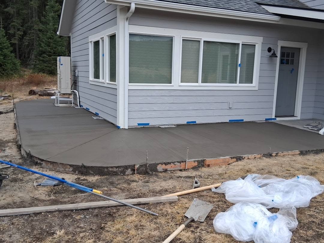 A fresh concrete patio surrounds a gray-sided house, with plastic sheeting, tools, and a blue hose scattered across the unfinished yard.