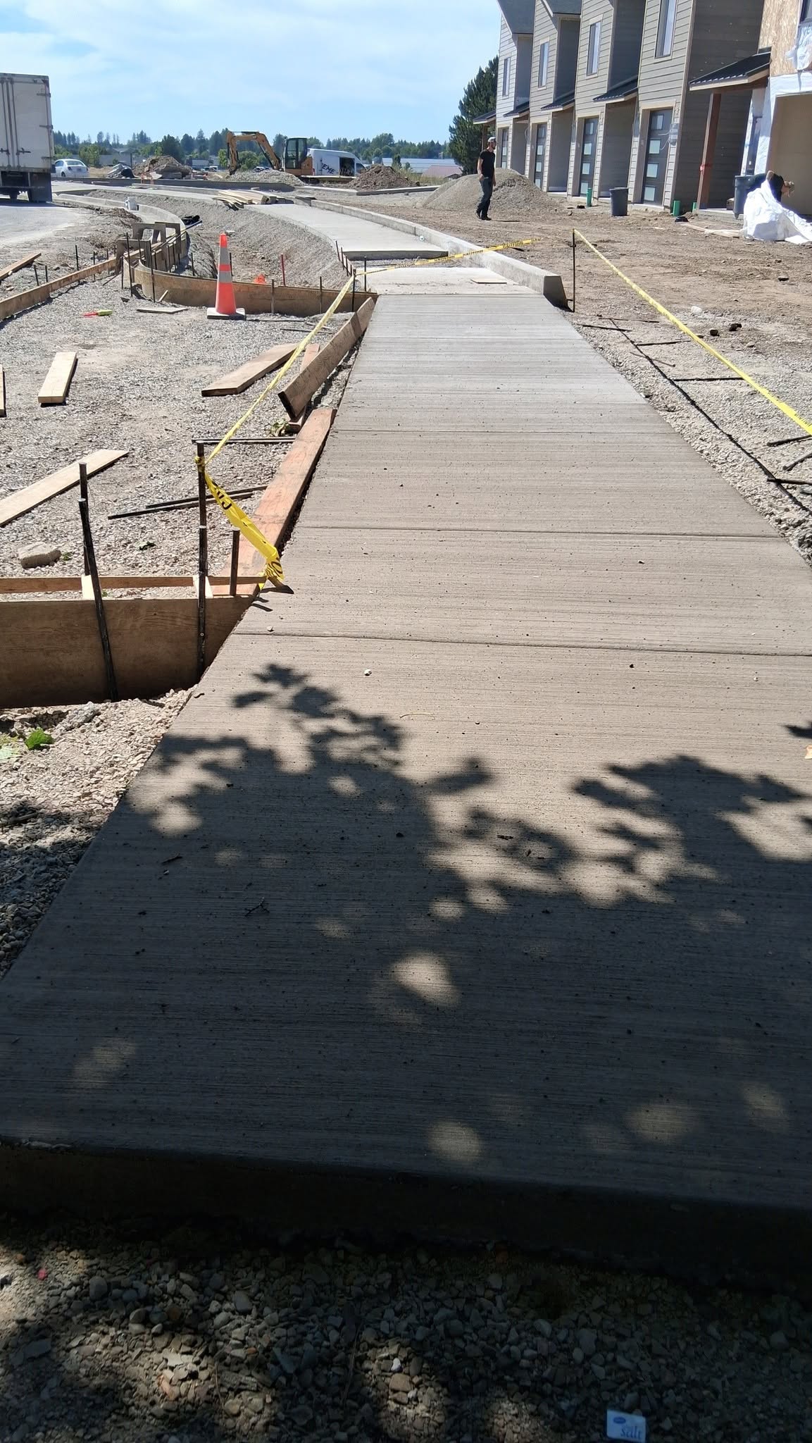 A new concrete sidewalk runs through an unfinished construction site beside townhouses, with trenches, cones, and caution tape under a clear sky.