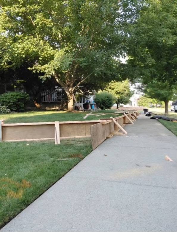 A quiet, tree-lined sidewalk borders a grassy yard enclosed by temporary wooden construction barriers, with tools and materials nearby on a sunny day.
