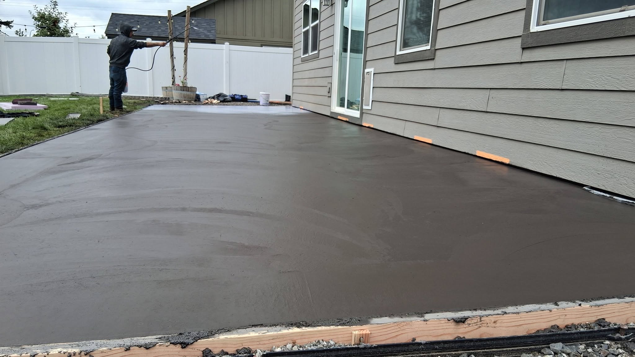 A worker smooths freshly poured concrete beside a gray-sided house, creating a wide, even patio in a quiet residential backyard.
