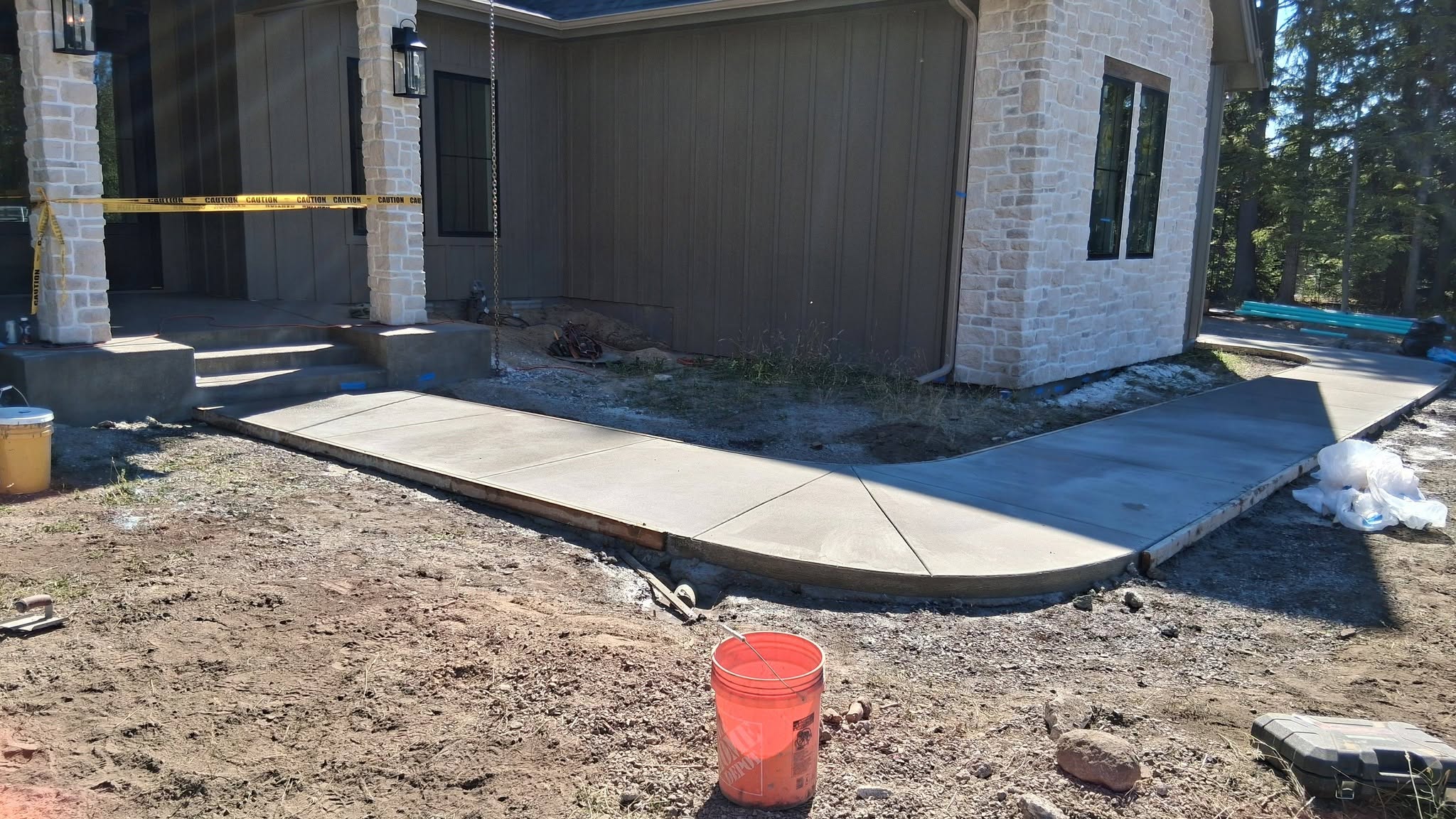 Fresh concrete walkway curves along a stone-and-dark-panel building under construction, with an orange bucket, exposed dirt, and caution tape conveying an active worksite.