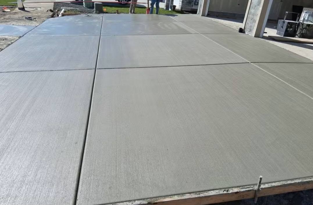 Freshly poured concrete driveway with broad, smooth slabs divided by straight expansion joints; workers and a garage appear in the background, conveying active construction.