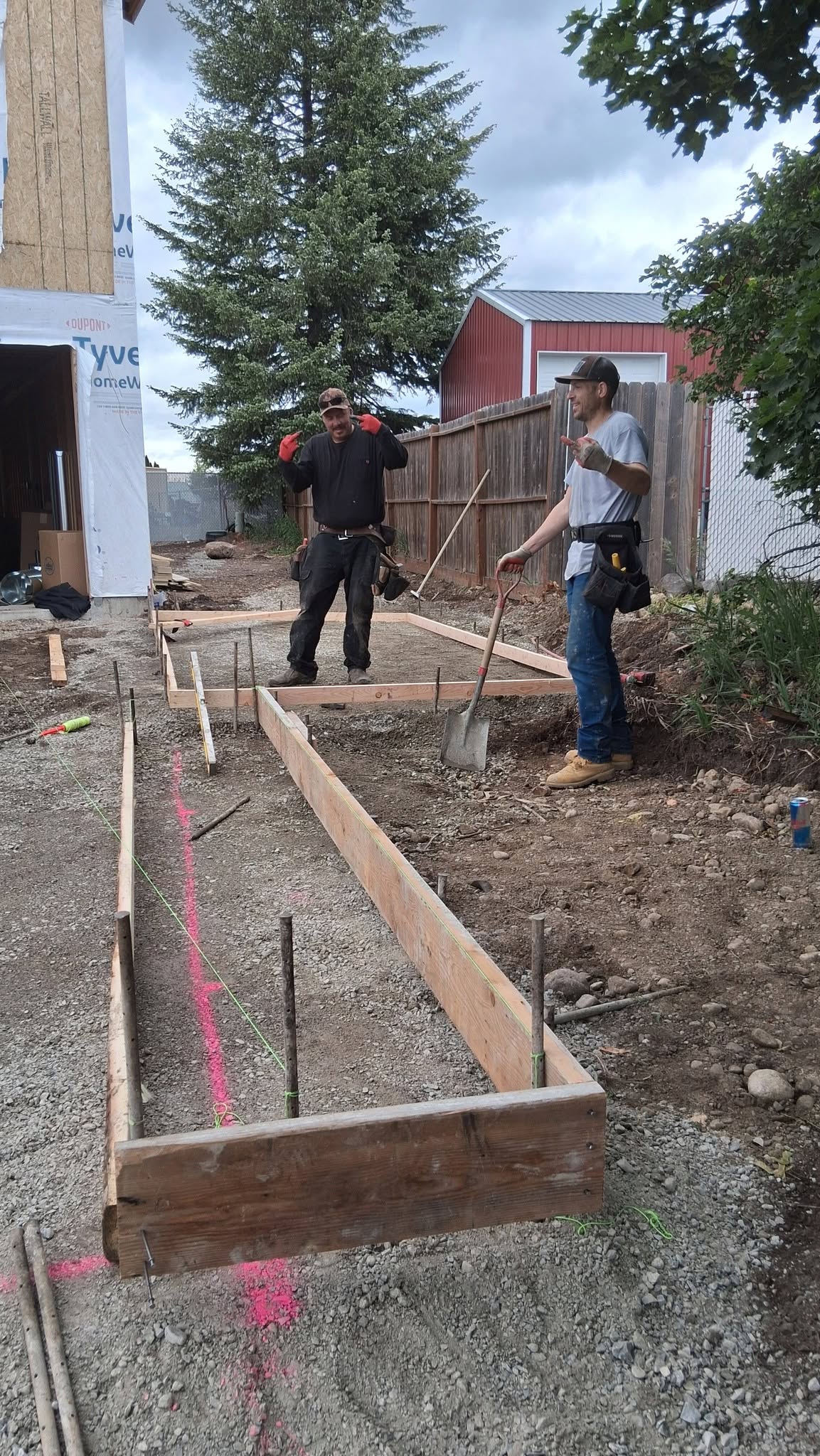 Two construction workers stand beside a wooden form for a walkway, with tools, pink layout markings, and a red barn in the background.